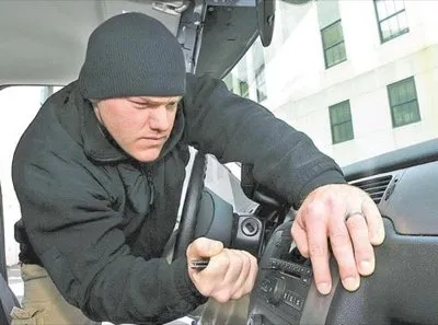 A person wearing dark clothing reaches through a broken car window, attempting to steal a car stereo system. The image illustrates vehicle break-in and theft.